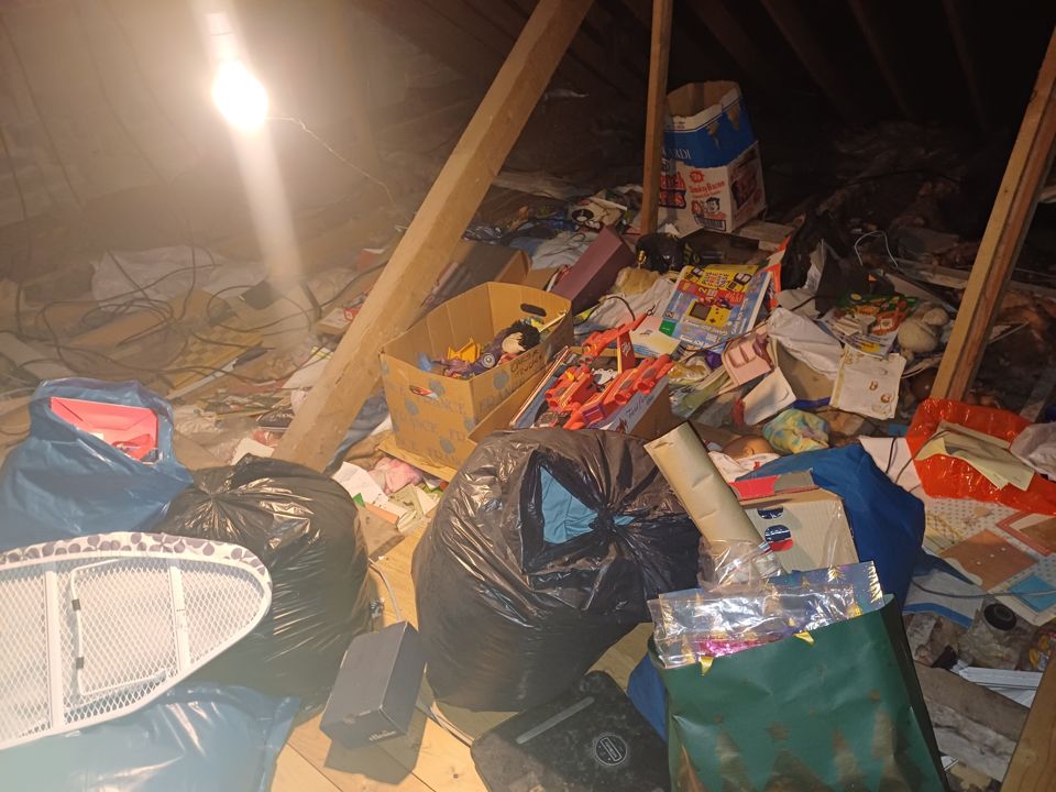 Attic hoard lit by a bare bulb, bin bags, boxes and toys piled between the roof timbers