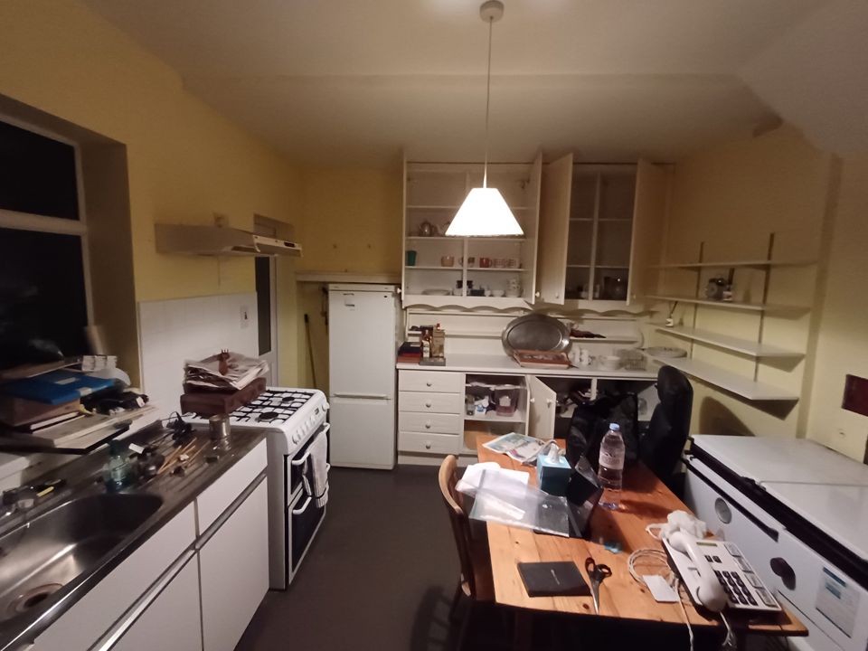 Dated kitchen before house clearance, cluttered worktops and table, open dresser cupboards, low evening light