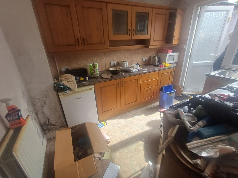 Old kitchen with damp walls, boxes and piled household items on worktops and floor