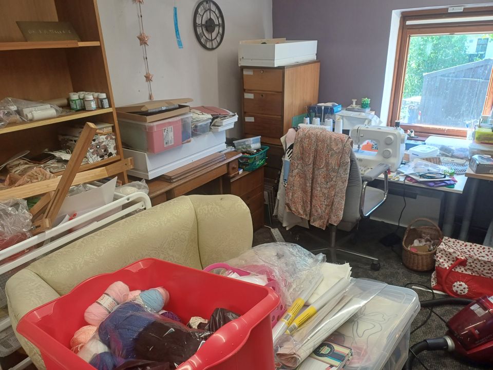 Cluttered craft room: armchair, wool baskets, shelves and desks piled high with stored items