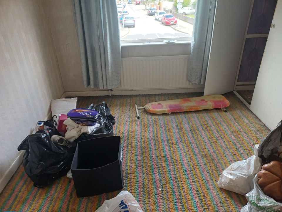 Bedroom mid-clearance with black bags, crates and loose belongings spread across patterned carpet