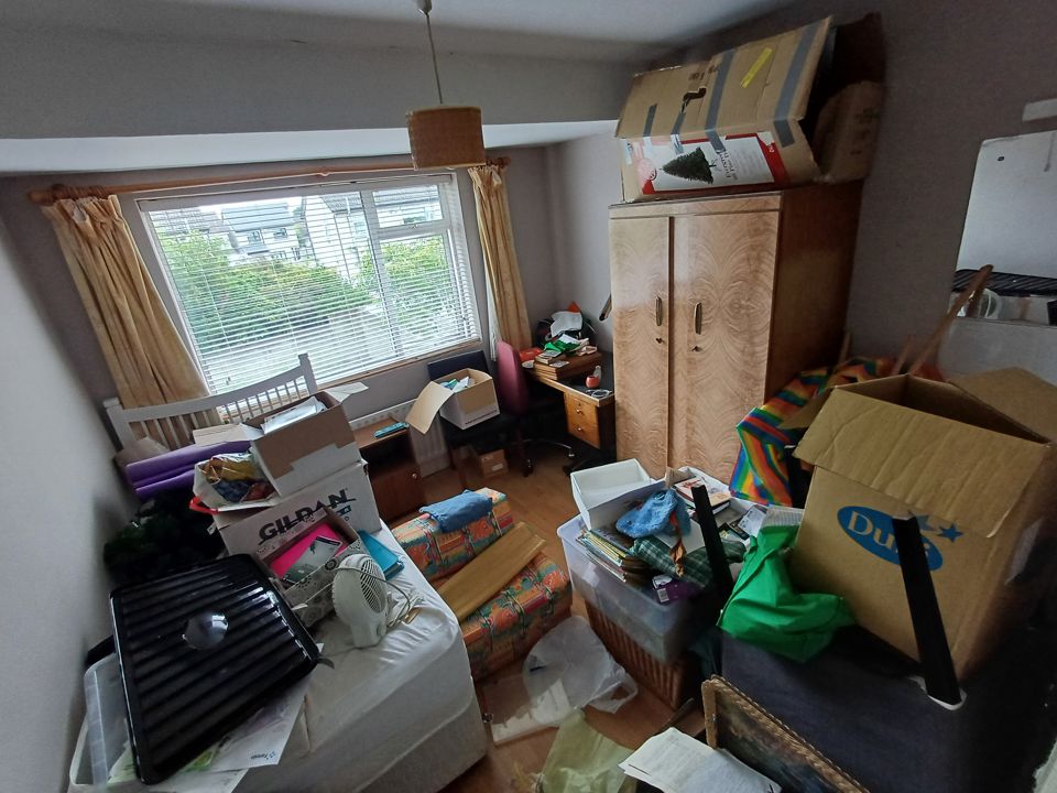 Bedroom packed with boxes, bags, fan heater, wardrobe topped with cartons; floor barely visible before clearance