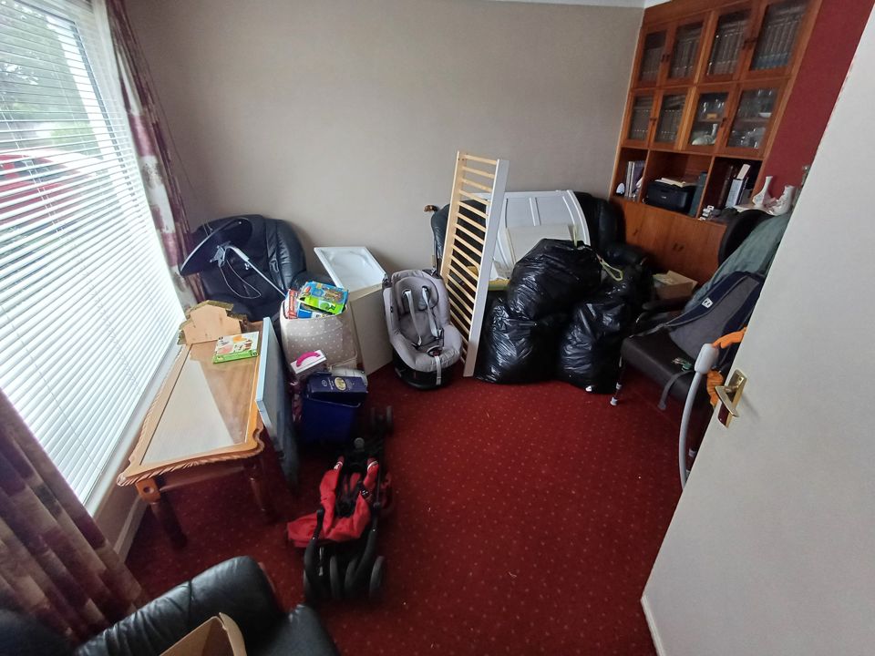 Front room on red carpet with bagged contents, cot frame, chairs and boxed items grouped for removal