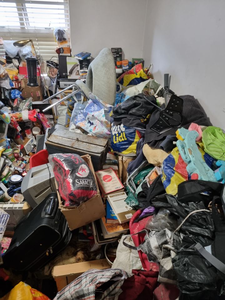 Same hoarded bedroom from another angle, waist-high piles of bags, clothes and boxes