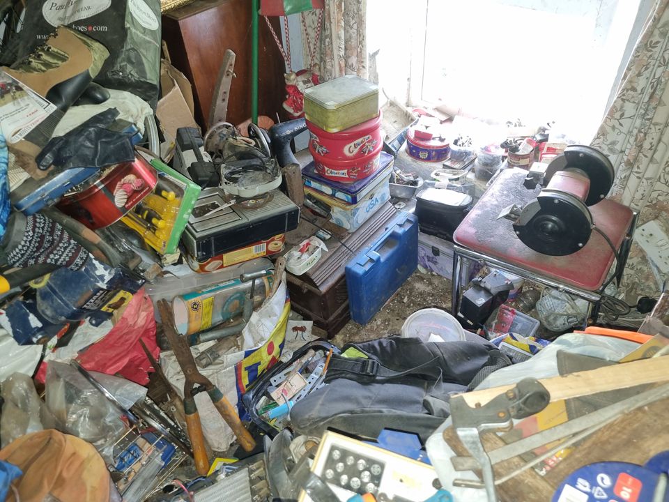 Floor-to-window clutter of tins, tools, bench grinder and bags in a hoarded room