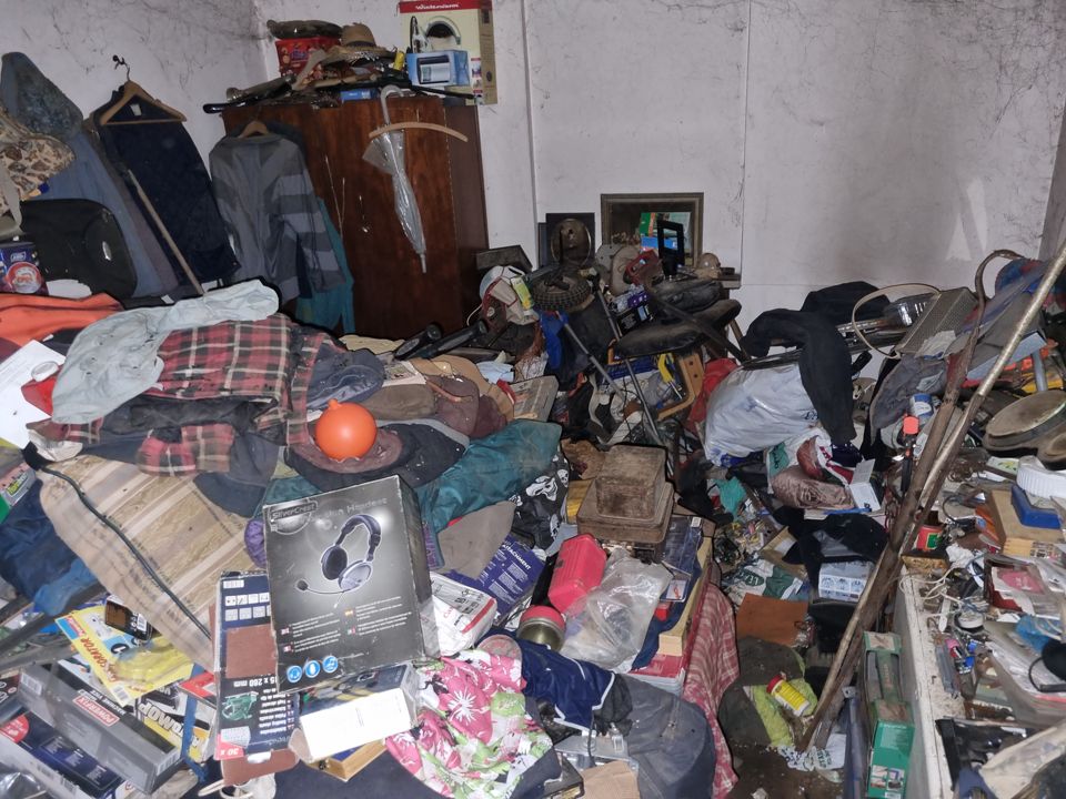Hoarded room packed with clothes, tools, boxes and junk piled high against wardrobe and walls