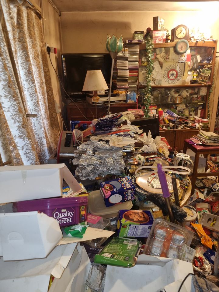 Hoarded living room with packaging, foil and clutter heaped in front of TV and cabinet