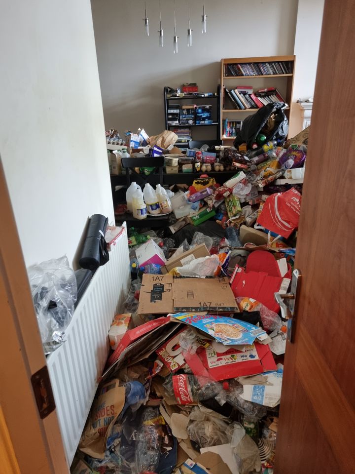 Hoarded dining room seen through doorway, floor buried knee-deep in packaging, bottles and boxes