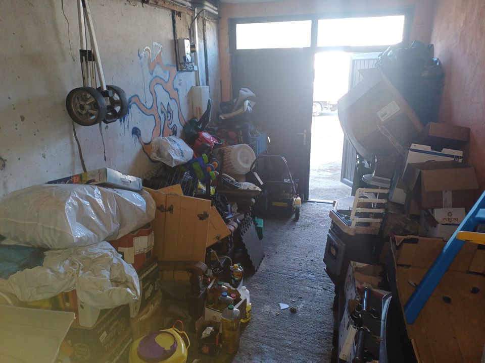 Long garage packed both sides with bags, boxes, furniture and toys, only a narrow path to the door