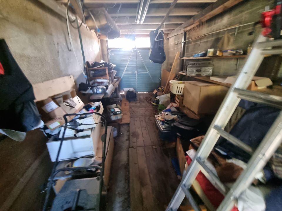 Long single garage crammed with boxes, ladder, lawnmower and shelving up to the door