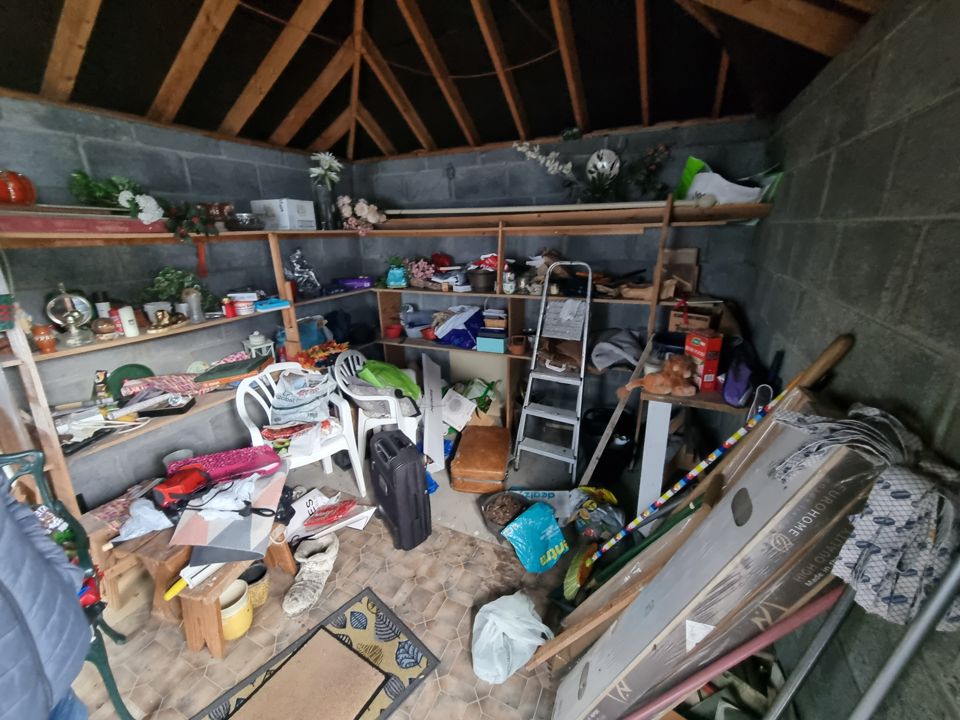 Block garage with shelves crammed with junk, chairs, suitcases, boards and bags on floor
