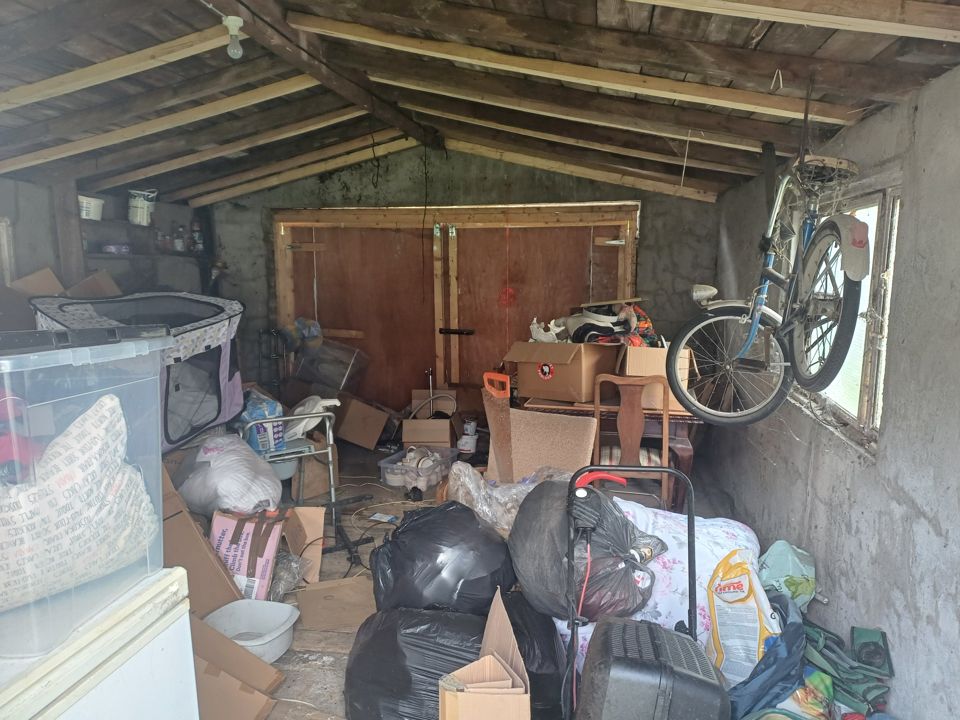 Old timber-roofed shed filled with bin bags, boxes, chairs, bicycle and discarded household items